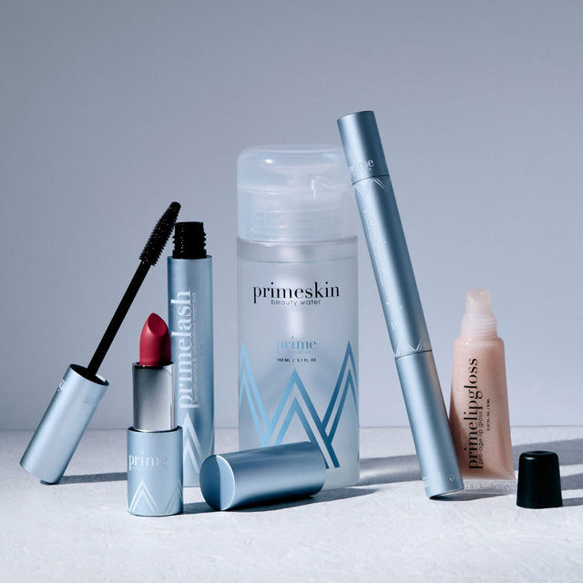 A set of Primeskin beauty products, including mascara, lipstick, primer, lip gloss, and a cleanser, arranged on a light surface with soft shadows.