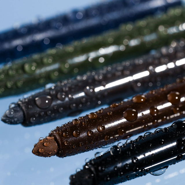 Close-up of five colorful eyeliner pencils with water droplets on them, arranged diagonally on a reflective surface. The pencils are blue, green, brown, black, and gray.