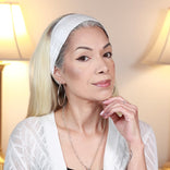 A woman with long blonde hair, wearing a white headband, white cardigan, and hoop earrings, poses indoors with her hand resting on her chin. Two lamps are visible in the softly lit background.