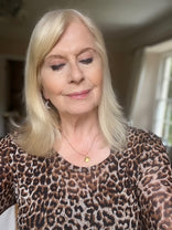 A woman with blonde hair and light makeup smiles gently with her eyes closed, wearing a leopard print top and a gold necklace, standing in a softly lit room with a window in the background.