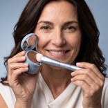 A smiling woman with wavy brown hair holds the PrimePrometics™ 4-in-1 Hairline Powder Brush, featuring cruelty-free bristles, as she prepares to apply powder to her face against a light blue background.