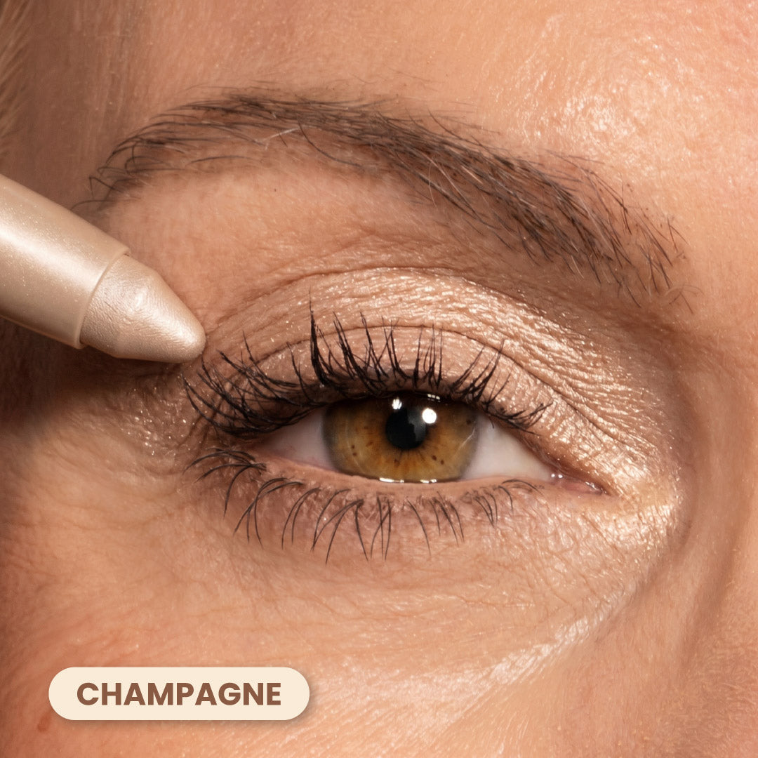 A close-up of an eye with champagne-colored eyeshadow applied to the eyelid. An eyeshadow pencil is being used on the upper lash line. The word “CHAMPAGNE” appears at the bottom left.