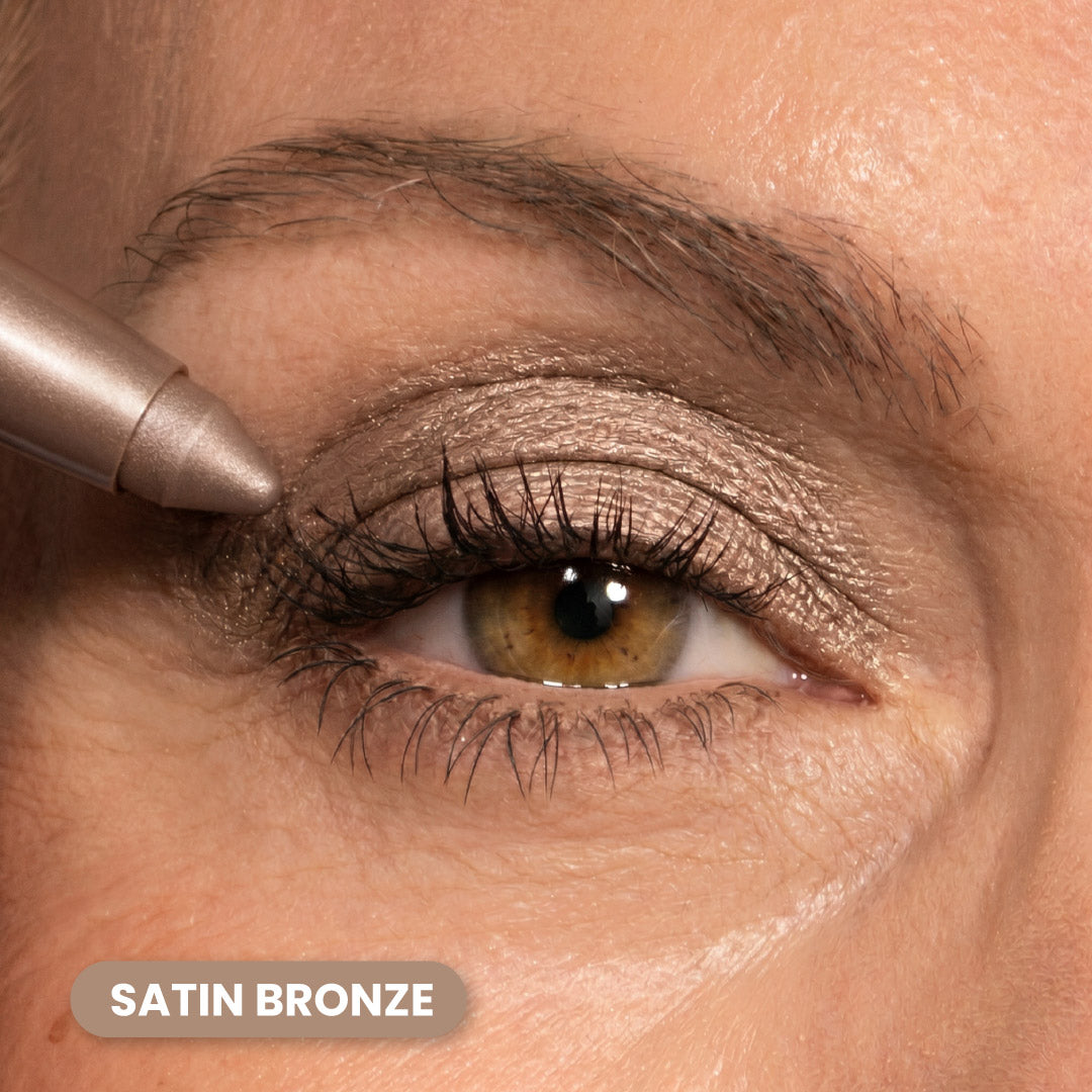 A close-up of a persons eye with bronze eyeshadow applied to the eyelid. An eyeshadow pencil is touching the outer corner. The text SATIN BRONZE appears at the bottom left of the image.
