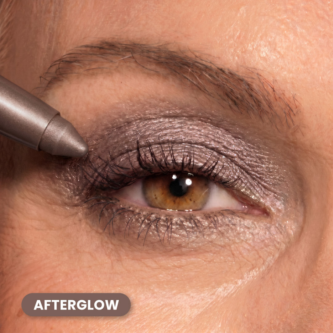 A close-up of a person’s eye with metallic purple eyeshadow being applied using a makeup pencil. The text “AFTERGLOW” appears in the bottom left corner.
