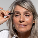 A smiling older woman with gray hair applies mascara to her eyelashes, holding a mascara wand near her right eye. She is wearing a white top and has natural makeup.