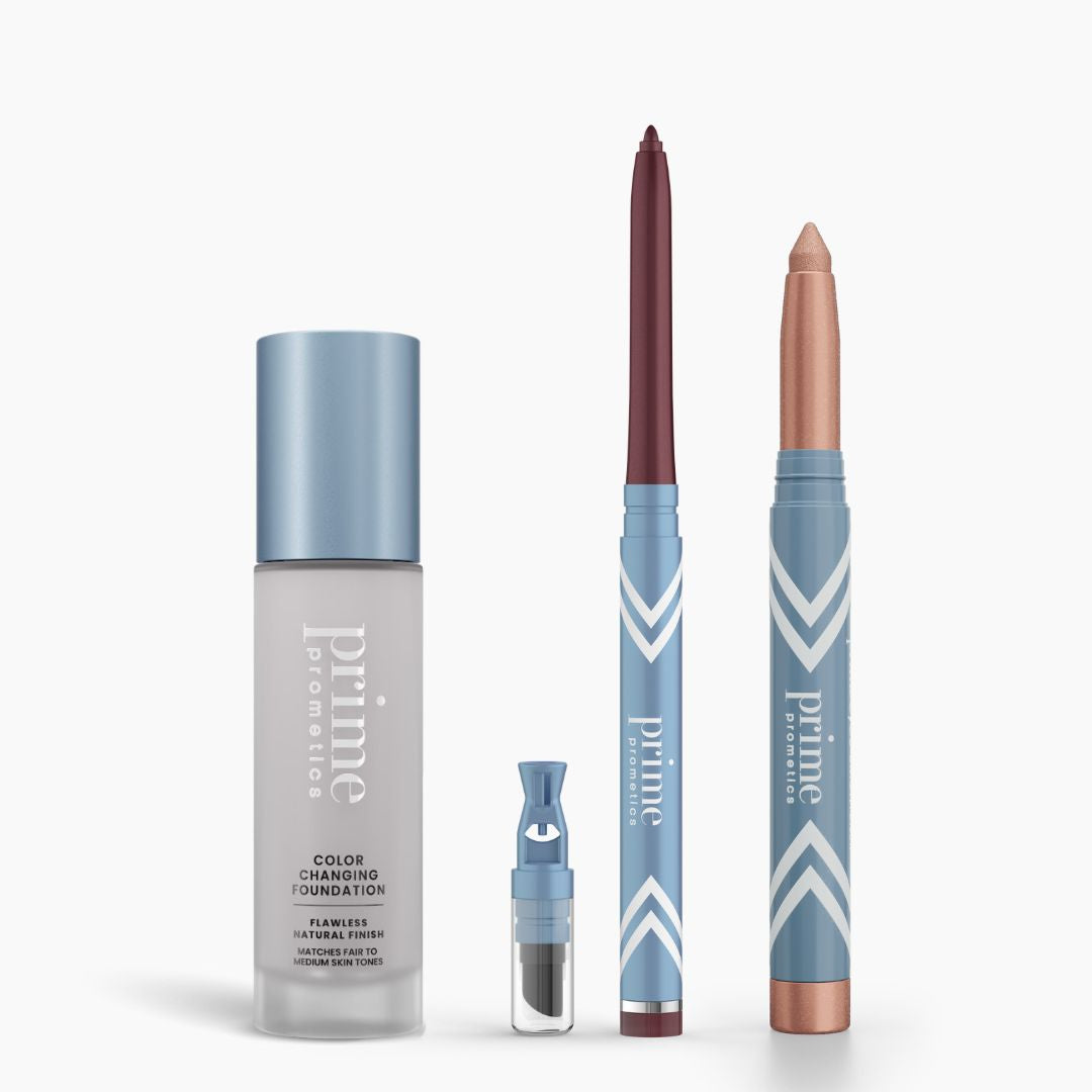 Four PrimePrometics™ All-Day Confidence Bundle products stand upright on a white background: a color-changing foundation, a small brightener vial, a maroon eyeliner pencil, and a shimmery beige eyeshadow stick in blue and white packaging.