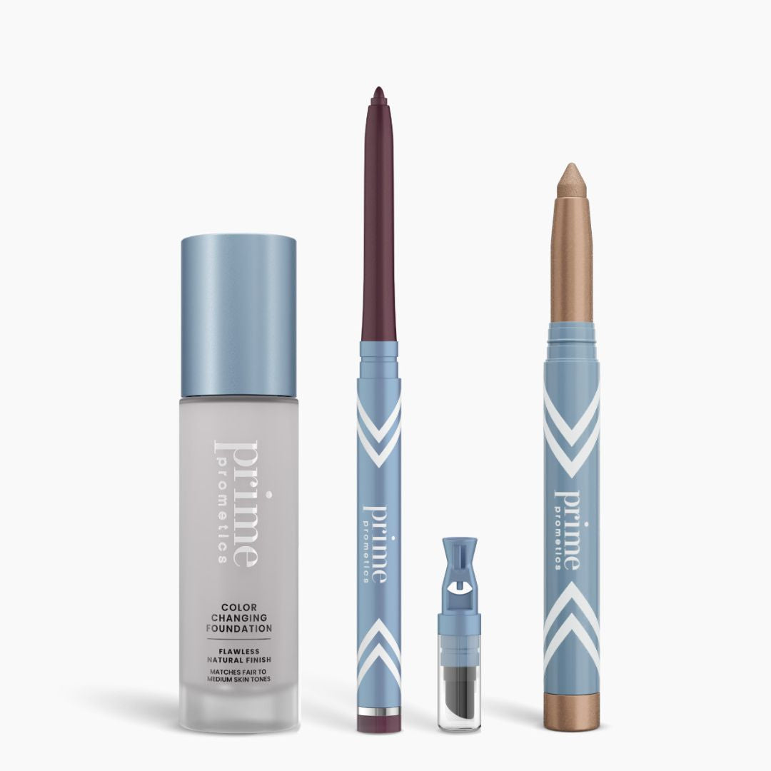 Four blue and silver PrimePrometics™ products from the All-Day Confidence Bundle—a color changing foundation, glide eyeliner pencil, brightener vial, and eyeshadow stick—are lined up against a white background.