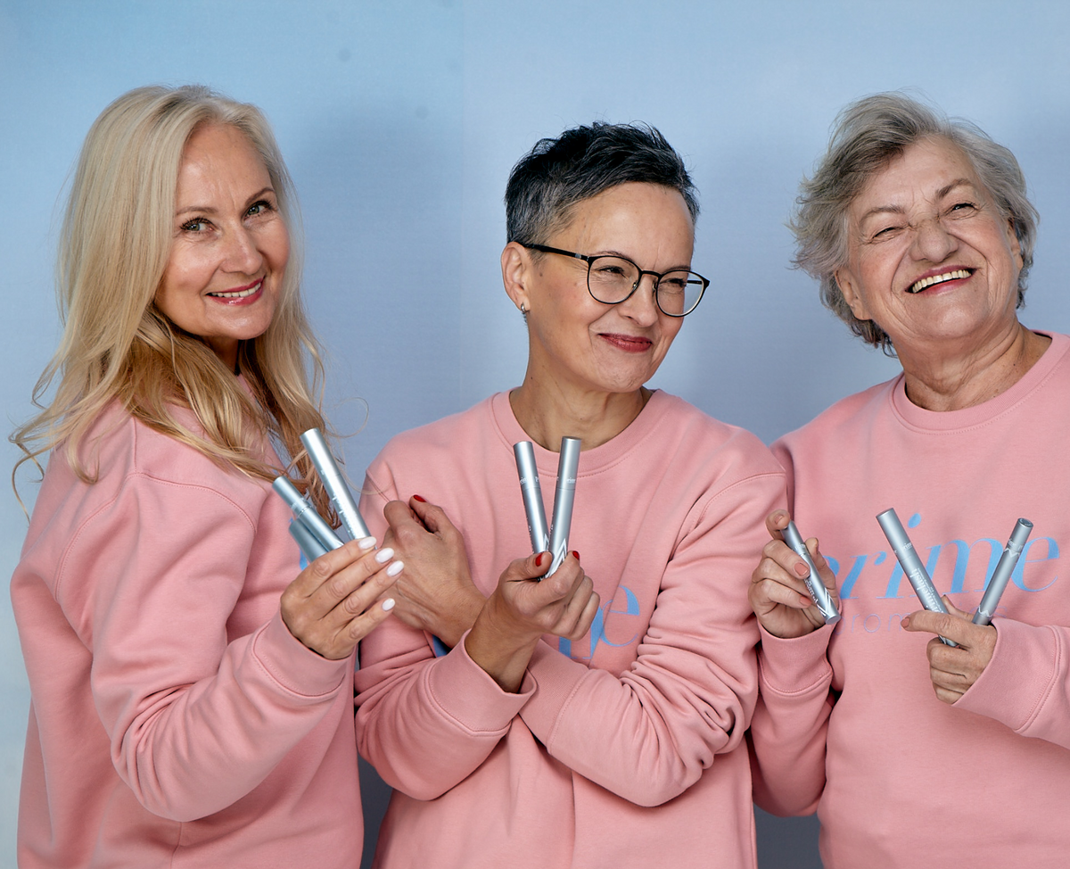 Three older women wearing matching pink sweatshirts smile while holding silver mascara tubes against a light blue background.