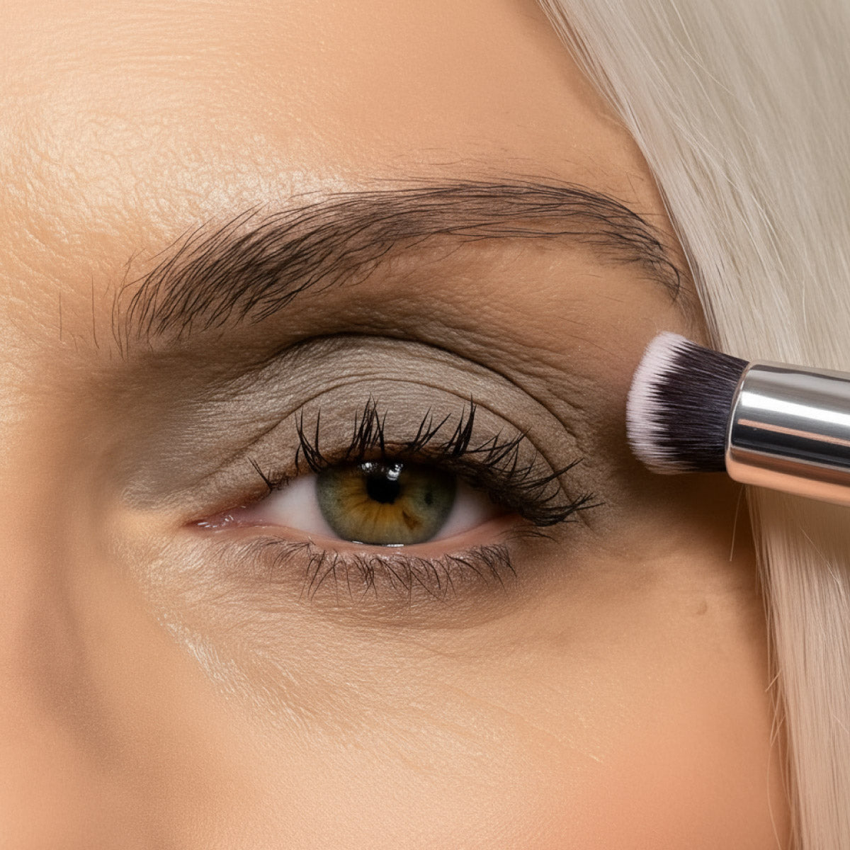 A close-up of a person with light skin and hazel eyes applying gray eyeshadow to their eyelid using a makeup brush. The persons eyebrow and part of their blonde hair are visible.