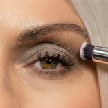 A close-up of a person with light skin and hazel eyes applying gray eyeshadow to their eyelid using a makeup brush. The persons eyebrow and part of their blonde hair are visible.