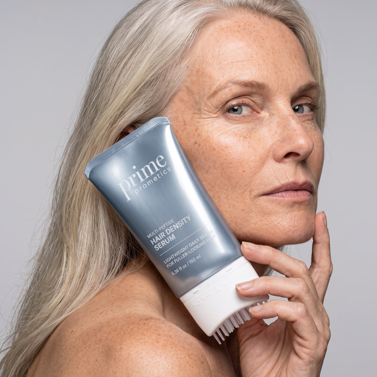 A woman with long gray hair holds a tube of PrimePrometics™ Multi-Peptide Hair Density Serum with a built-in applicator brush near her face, showcasing the product for fuller-looking hair.