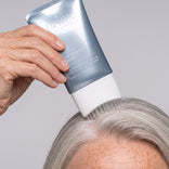 A hand applies PrimePrometics™ Multi-Peptide Hair Density Serum from a silver tube with a brush tip to the scalp of a person with gray hair, helping support fuller-looking hair.