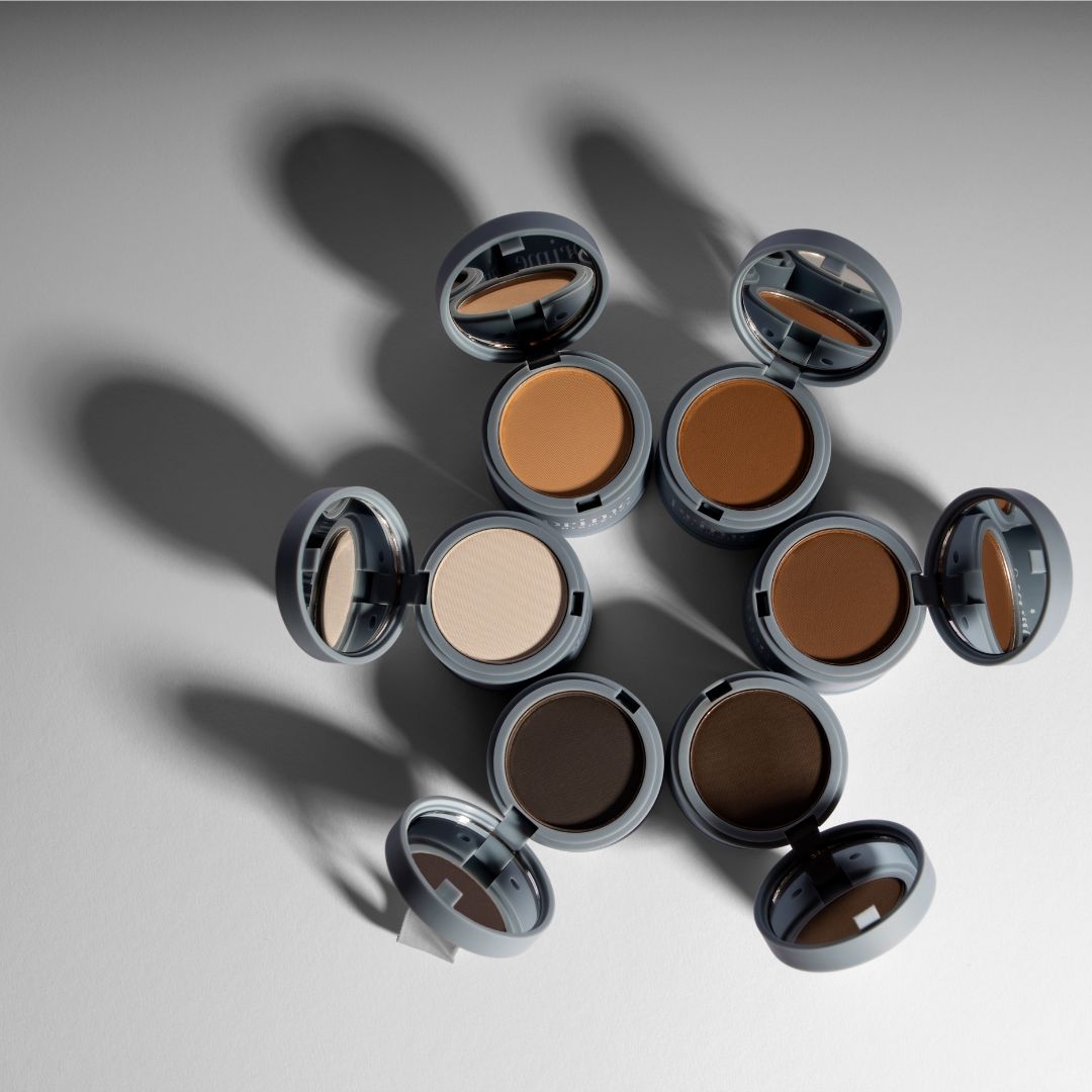 An overhead view of seven open compact powders in different shades, arranged in a circular pattern on a light surface with shadows cast behind them.