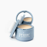 An open, round light blue compact labeled PrimePrometics™ Instant Coverage Hairline Powder reveals a mirror, beige powder for gray root coverage, and a white applicator puff on the side against a white background.