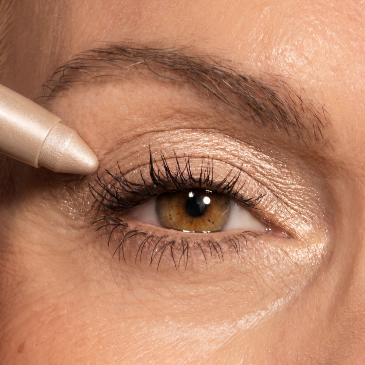 A close-up of an eye with brown and gold eyeshadow. An eye pencil is being applied to the outer corner of the upper eyelid. Eyebrow and skin details are visible.