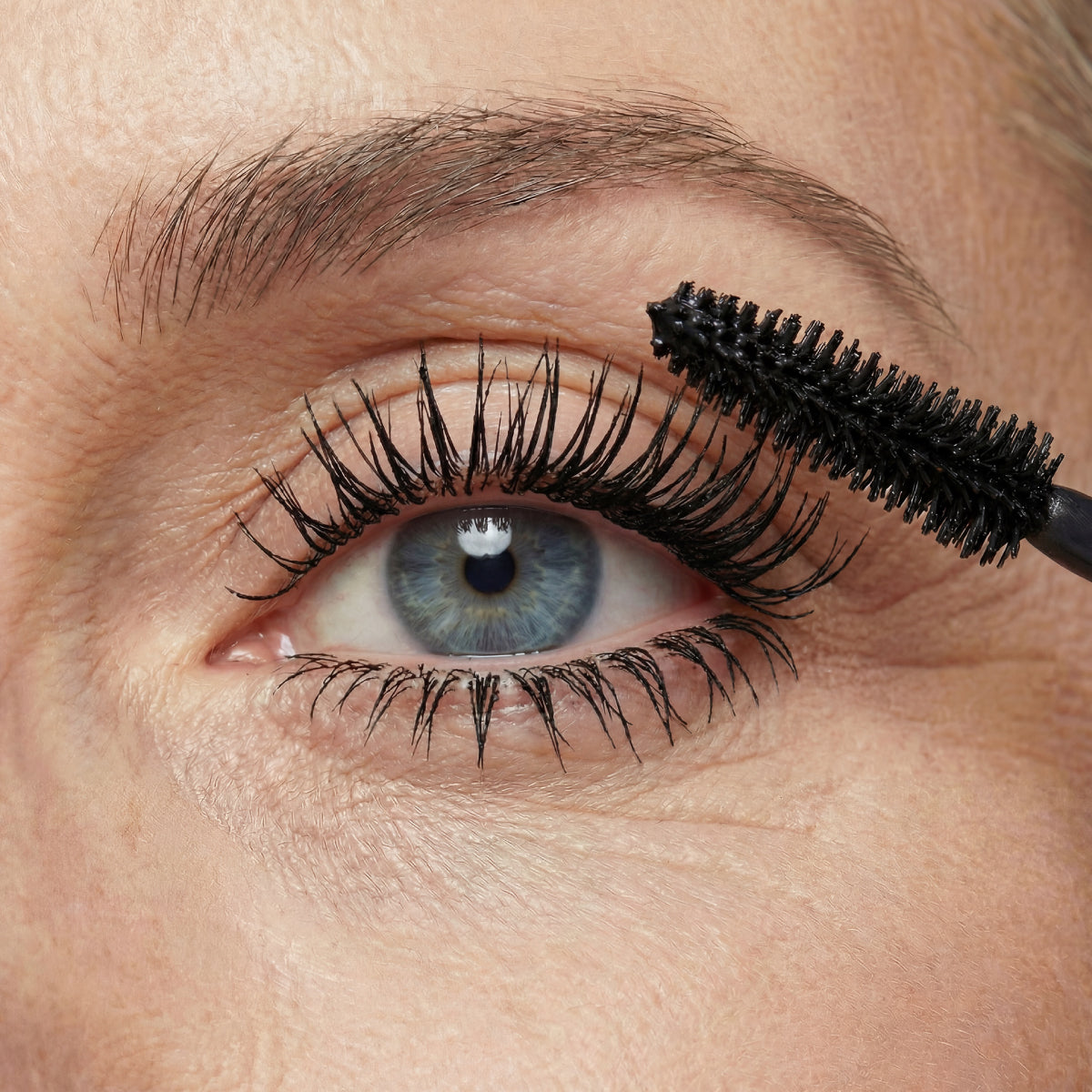 A close-up of a blue eye with mascara being applied to the upper eyelashes using a black mascara wand. The skin around the eye shows natural texture and fine lines.