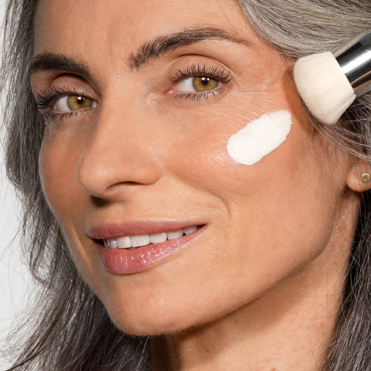 A woman with gray hair smiles while applying white cream to her cheek using a makeup brush. The image is focused on her face and the product being applied.