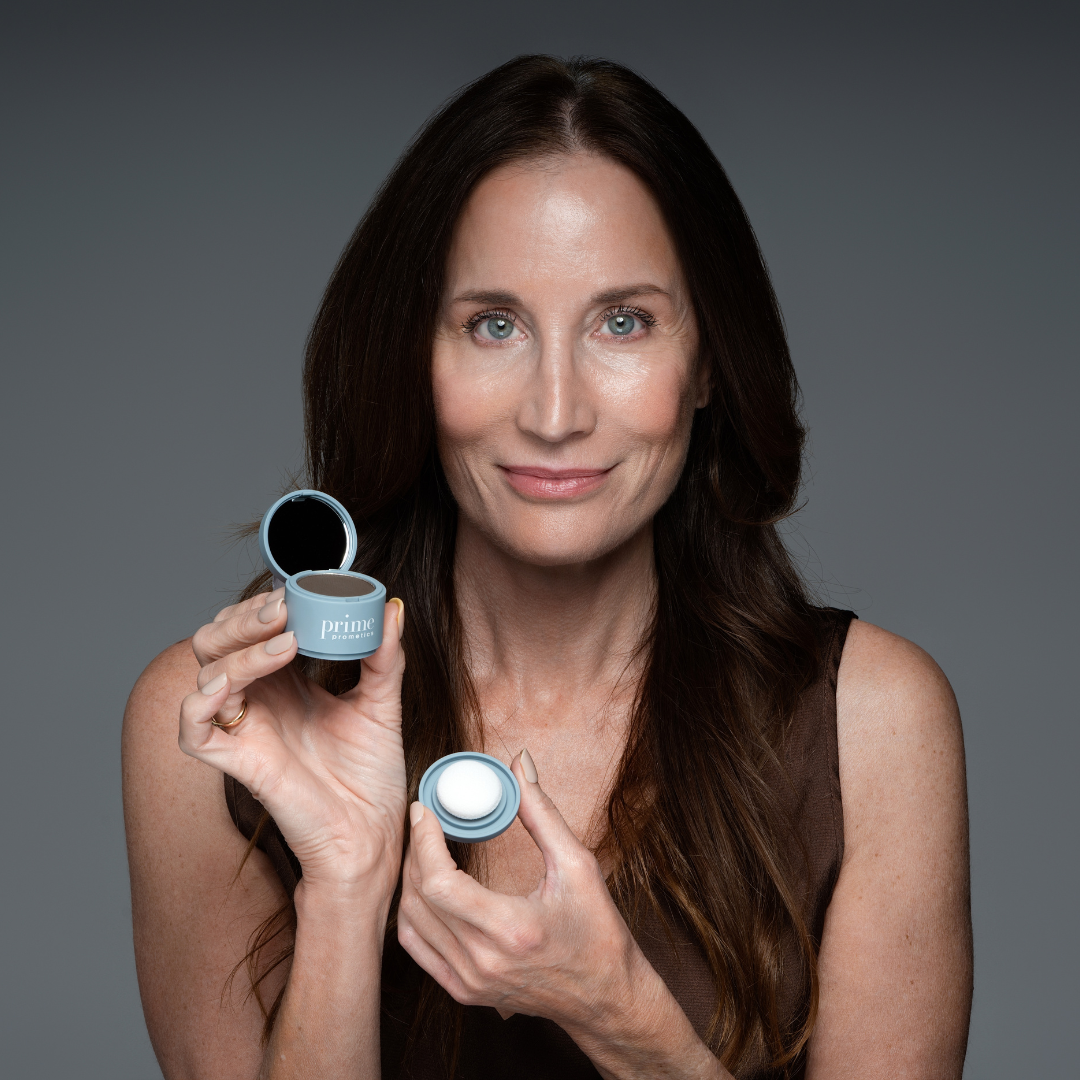 A woman with long brown hair holds an open compact makeup case with a mirror in one hand and the lid in the other, smiling softly against a neutral gray background.