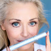 Mature blonde woman holding the Prime Prometics PrimeLash Mascara tube near her face, lashes visibly defined