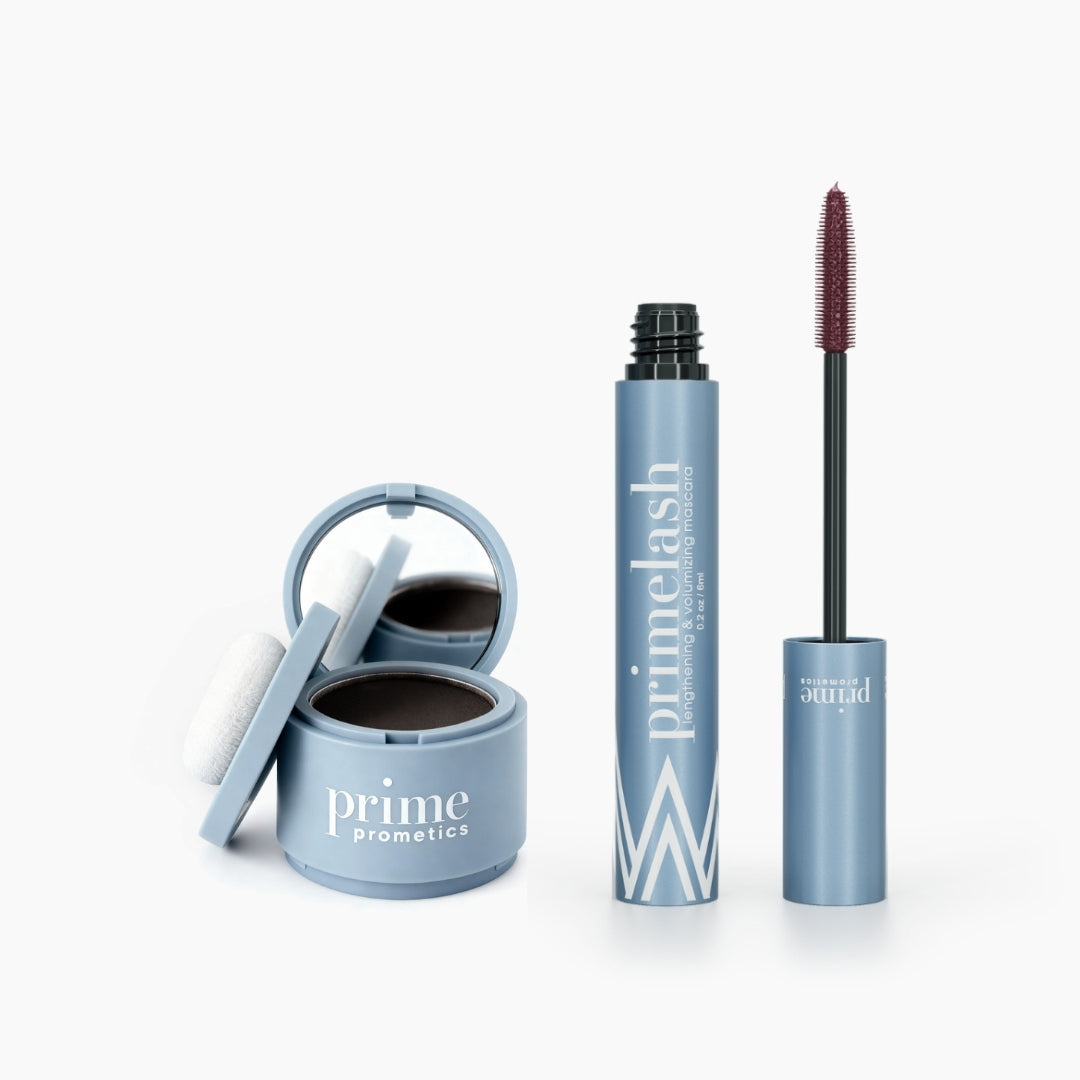 The PrimePrometics™ Beauty Essentials set features a light blue hairline powder compact with mirror and white puff beside a matching PrimeLash mascara tube with its exposed wand, all displayed against a clean white background.