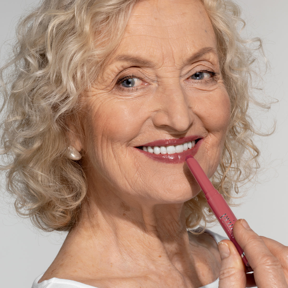 Smiling older woman with curly gray hair applies pink lipstick to her lips. She has blue eyes, wears pearl earrings, and is dressed in a white top against a plain light background.