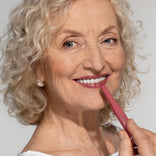 Smiling older woman with curly gray hair applies pink lipstick to her lips. She has blue eyes, wears pearl earrings, and is dressed in a white top against a plain light background.