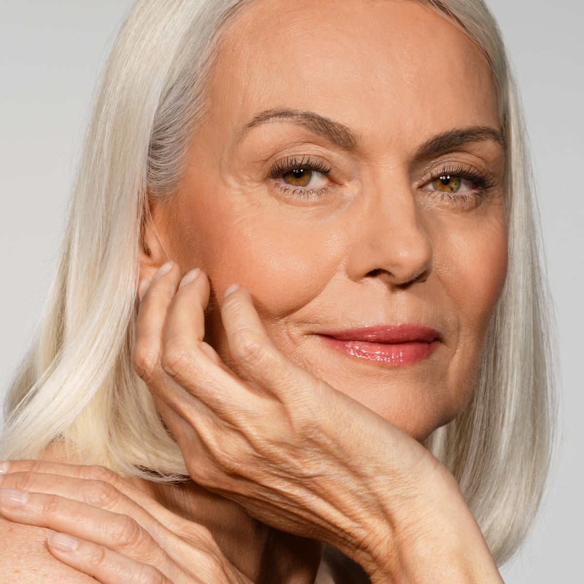 Close-up of an older woman with smooth, silver hair and glowing skin, gently resting her chin on her hand and smiling softly.