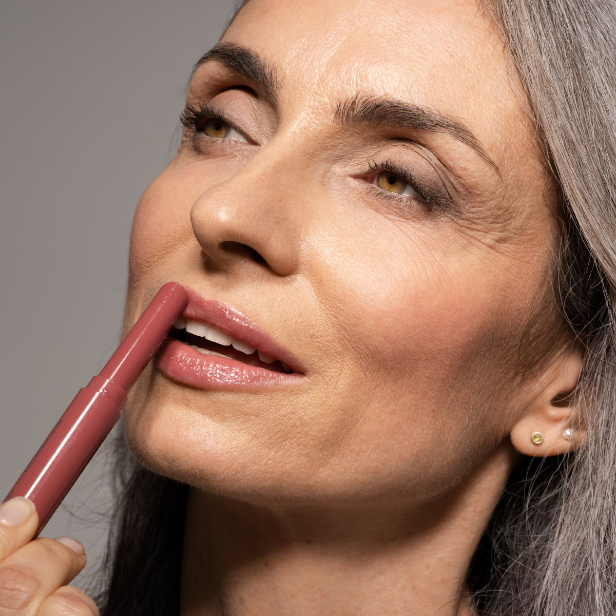 A woman with gray hair applies pink lipstick, smiling softly. She has light makeup and glowy skin, with stud earrings visible on her ear. The background is neutral and her expression is calm and content.