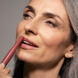 A woman with gray hair applies pink lipstick, smiling softly. She has light makeup and glowy skin, with stud earrings visible on her ear. The background is neutral and her expression is calm and content.