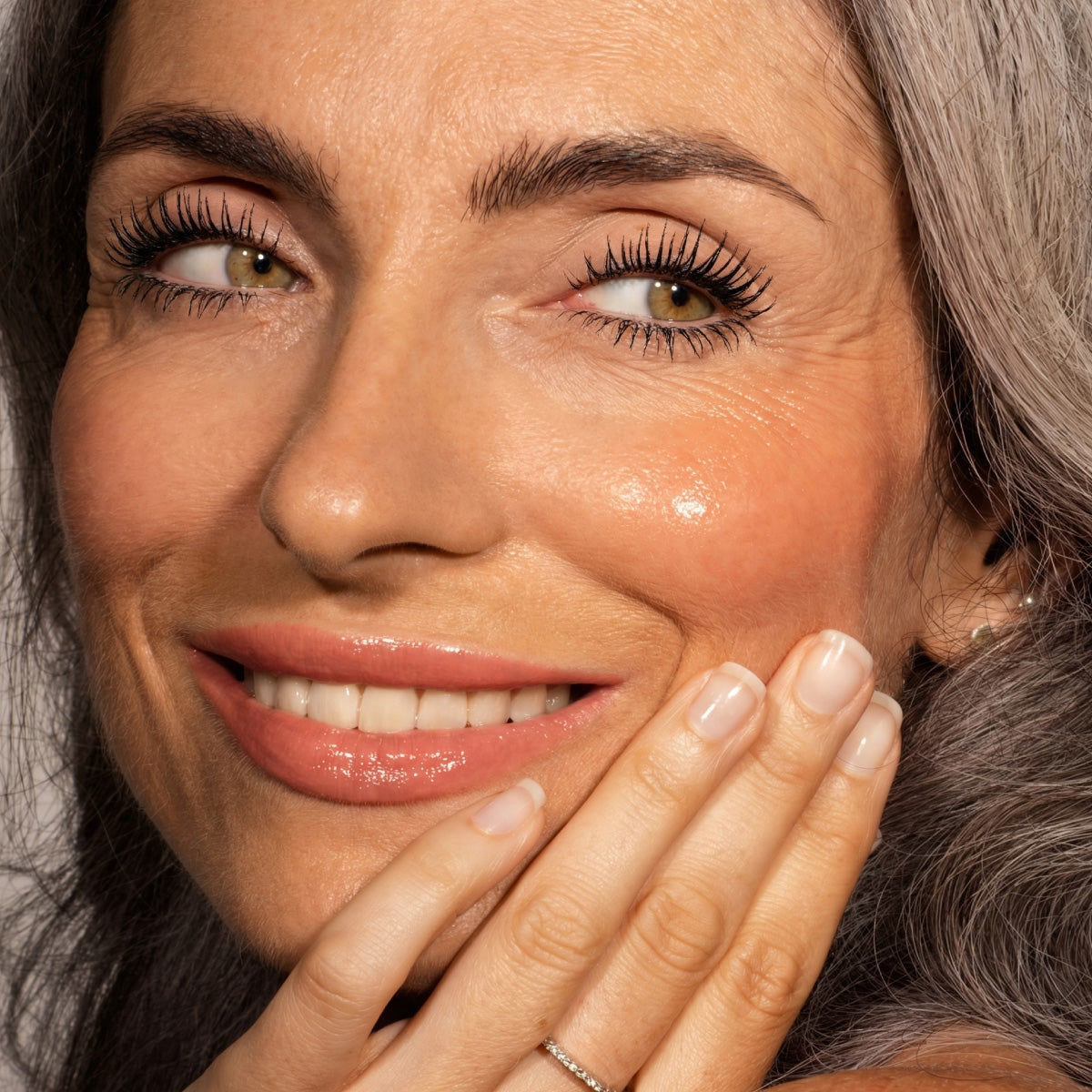 A close-up of a smiling woman with long gray hair and natural makeup, touching her face to show off manicured nails and a thin ring. She wears the PrimePrometics™ Instant Red Carpet Look Set.