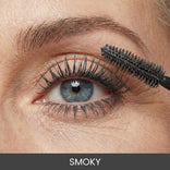 A close-up of a blue eye with long, dark eyelashes, as a mascara wand is applied to the upper lashes. The word SMOKY appears at the bottom of the image.