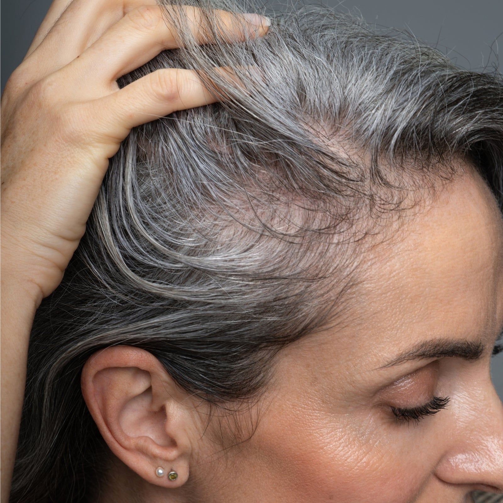A close-up of a woman with gray hair pulling her hair back to reveal thinning hair along her scalp and hairline. Her skin appears smooth and she is wearing small stud earrings.