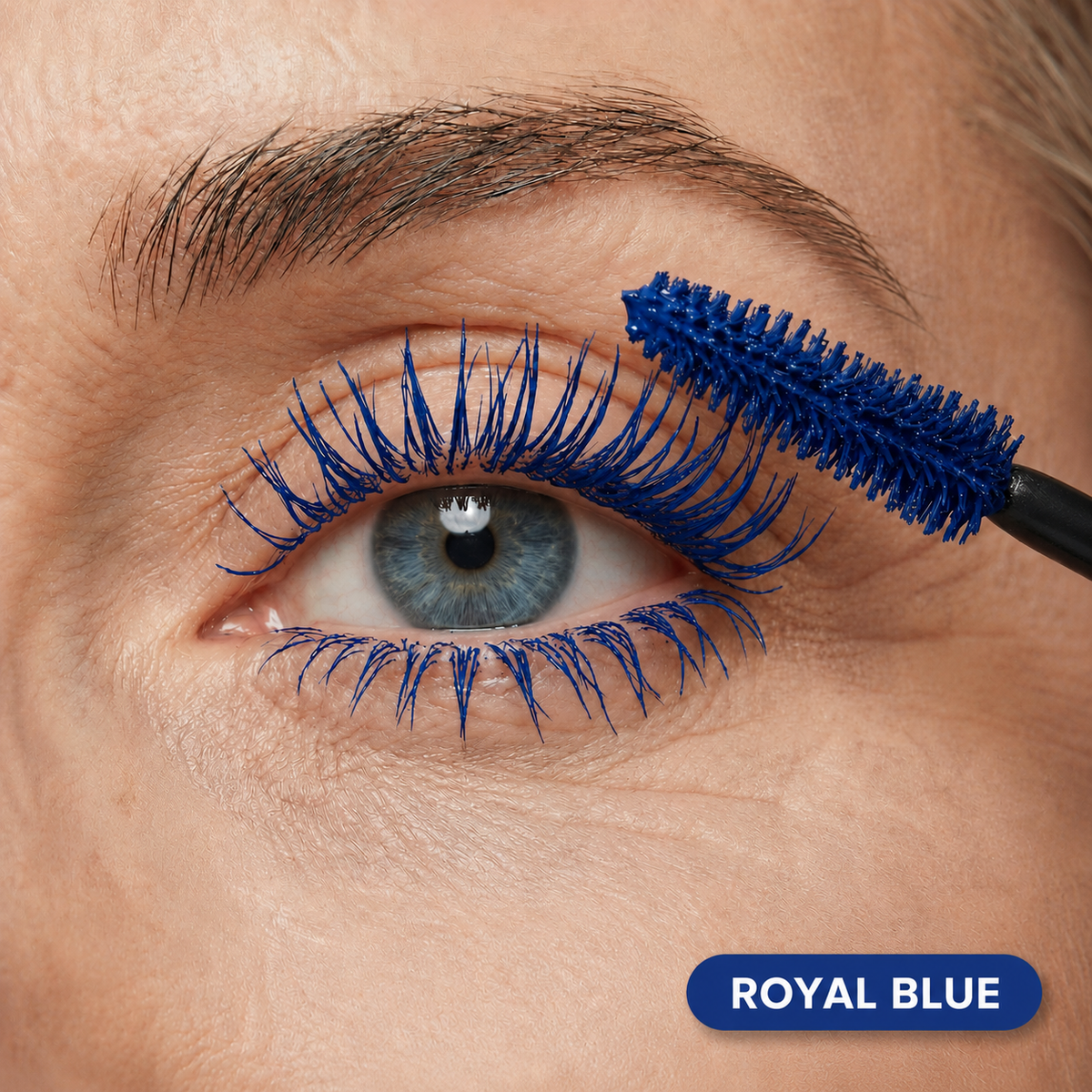 A close-up of an eye with PrimePrometics™ PrimeLash Mascara in royal blue being applied to the upper lashes. The mascara wand is visible, and a label in the lower right corner reads “ROYAL BLUE.” Specially formulated for mature women, this pro-age mascara boosts lash growth while delivering a bold, vibrant color.