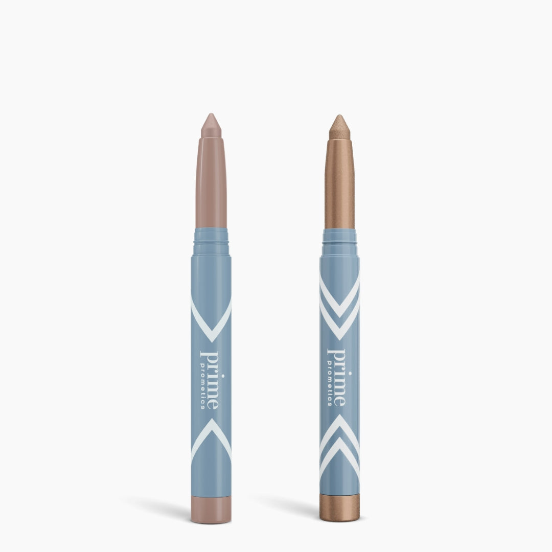Two blue PrimePrometics™ Complete Eye Sticks stand upright—one nude matte, one shimmery bronze. Both feature angled tips and minimalist design, making them the perfect everyday eye brighteners.