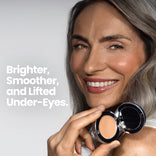 Smiling woman with long gray hair holds an open compact of PrimePrometics™ Undereye Brightening Corrector, revealing radiant skin. Text on image: Brighter, Smoother, Lifted Under-Eyes—ideal for reducing dark circles.
