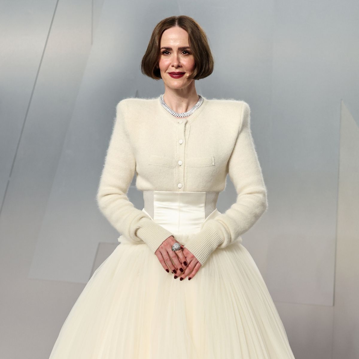 A woman with a bob hairstyle wears an elegant cream gown with a fitted bodice and full tulle skirt, accessorized with jewelry and dark nail polish. She completes her style using the Instant Red Carpet Look Set by PrimePrometics™.