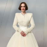 A woman with a bob hairstyle wears an elegant cream gown with a fitted bodice and full tulle skirt, accessorized with jewelry and dark nail polish. She completes her style using the Instant Red Carpet Look Set by PrimePrometics™.