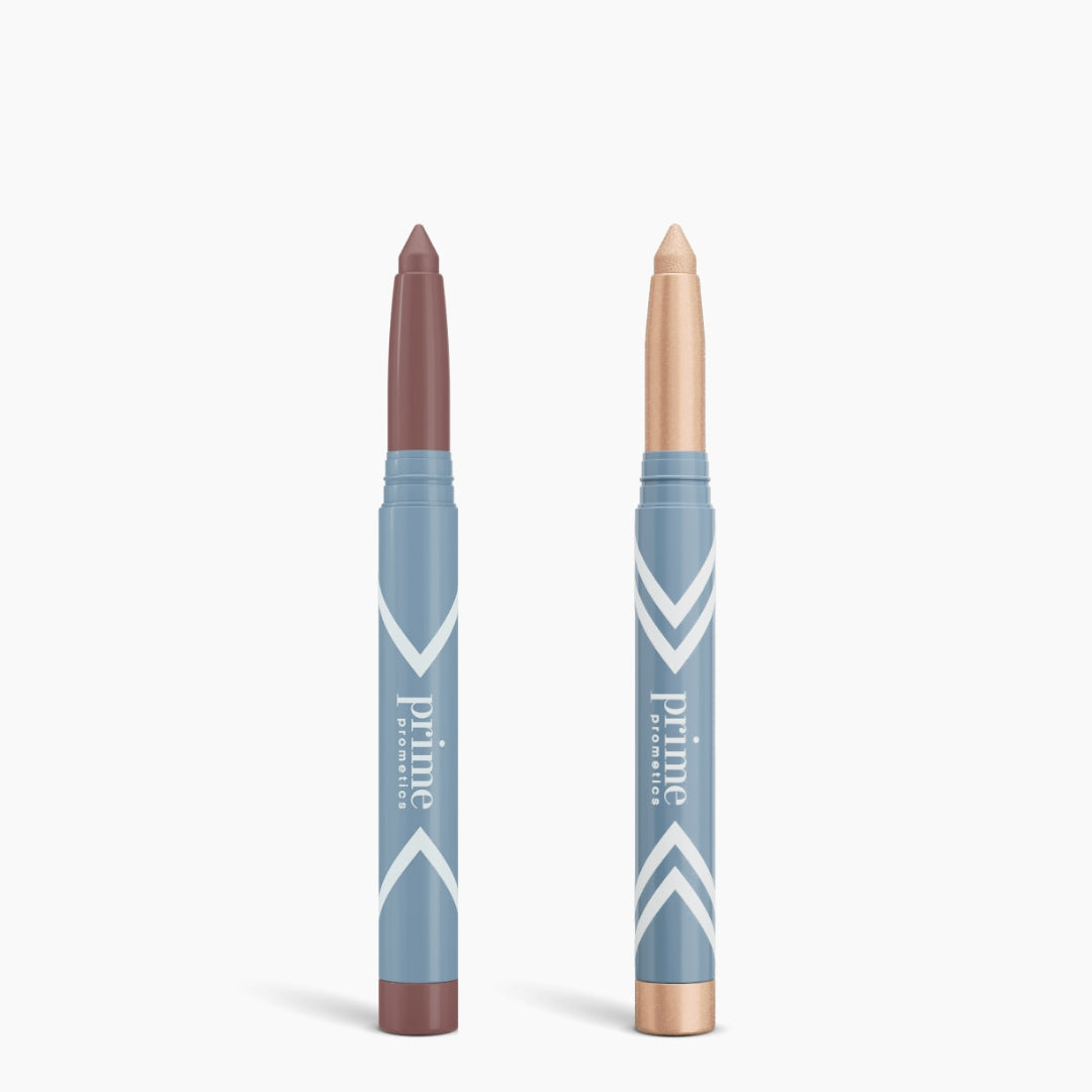 Two blue PrimePrometics™ Complete Eye Sticks Kit tubes stand upright. The left offers mauve matte eyeshadow; the right is a shimmery champagne eye brightener. Both feature twist-up packaging and white chevron patterns.